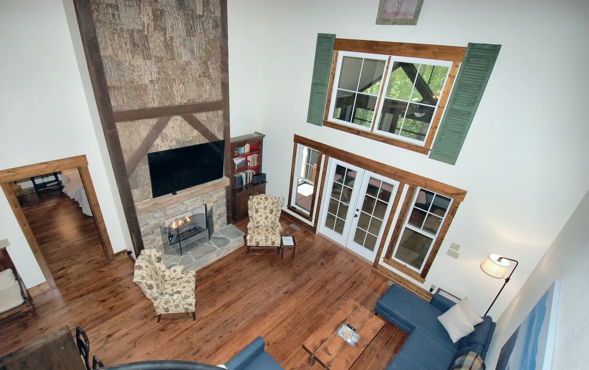 The image showcases a cozy, inviting lodge interior featuring a stunning stone fireplace, plush seating area, exposed wooden beams, and expansive windows that frame lush, woodsy views. The warm glow of the fire, natural textures, and abundant natural light set the scene for relaxation and connection.