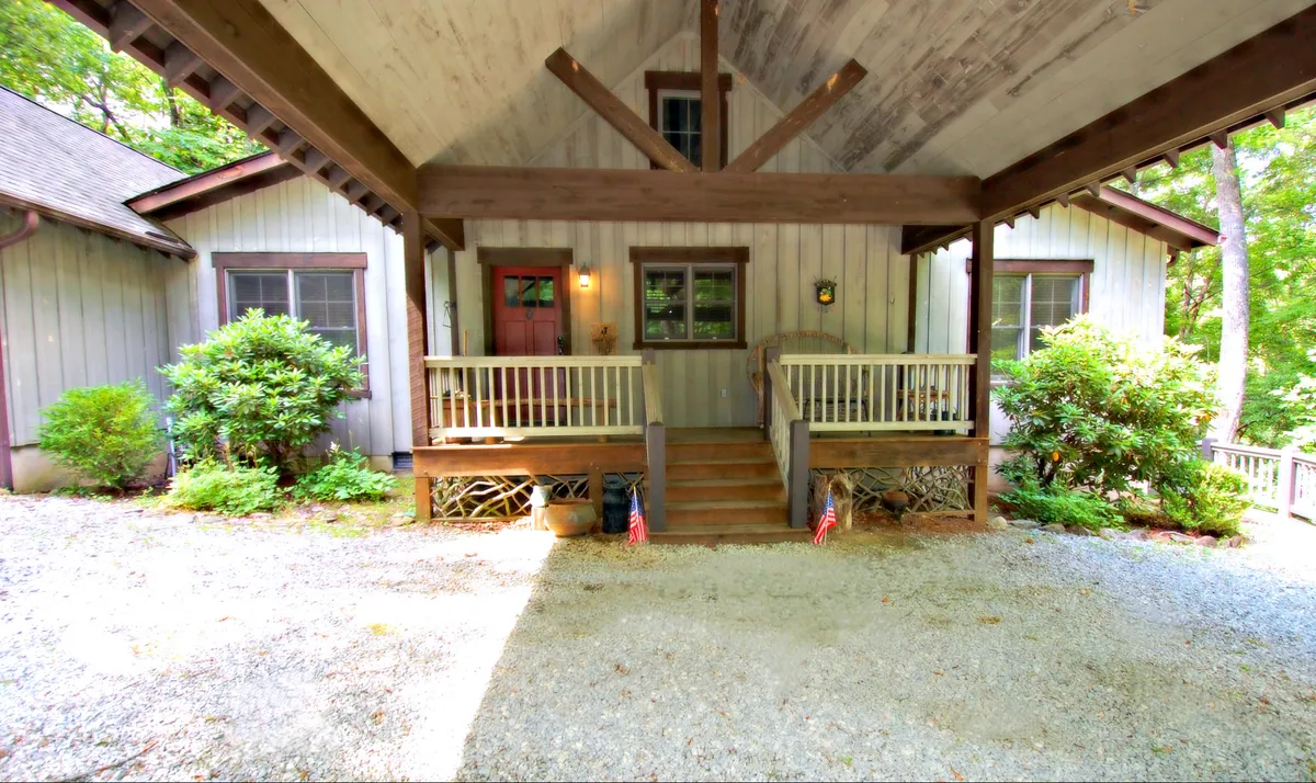 This exterior image shows a welcoming wooden porch at the front of the property, with decorative flag bunting and green shrubs flanking the entrance. The porch is furnished for relaxation and socializing, suggesting a friendly, homey atmosphere that invites guests to gather and enjoy the outdoors. The scene hints at the connection between the home's rustic charm and the vibrant town nearby.