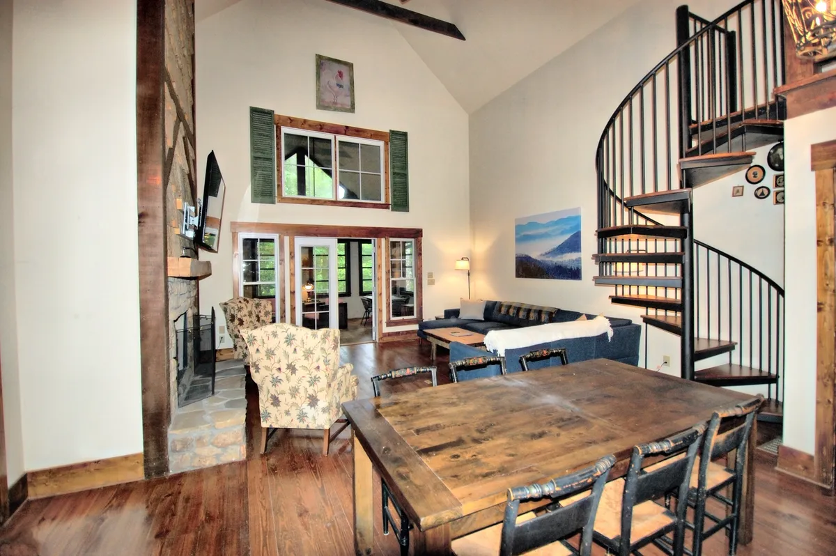 A spacious living area featuring a captivating stone fireplace as its focal point, with a spiral staircase adding rustic character. Large windows bathe the seating area and dining table in natural light, creating an inviting atmosphere for gathering and relaxing.