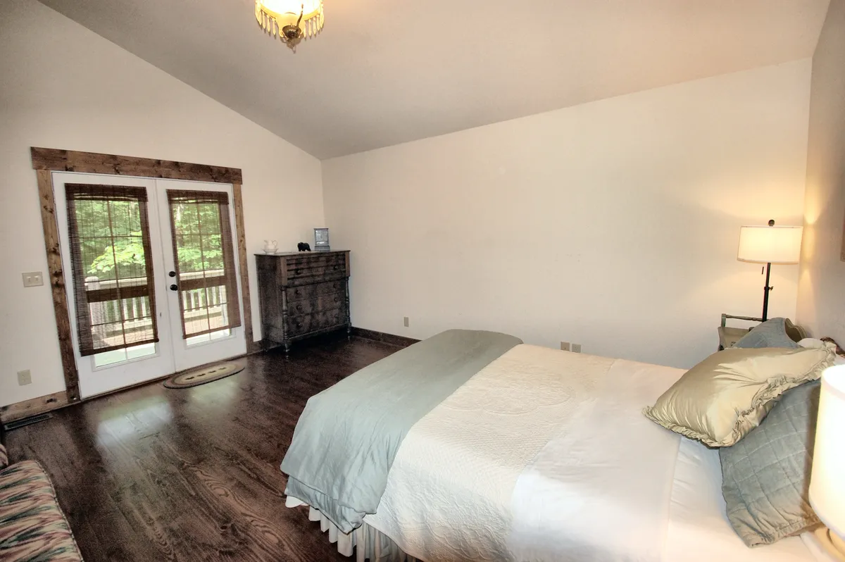 A peaceful bedroom scene featuring a bed with crisp linens, a dresser topped with decorative objects, and double doors that open onto a balcony with a glimpse of the natural surroundings beyond. Soft lighting and nature-inspired décor evoke tranquility. The presence of the balcony hints at an easy transition from indoor comfort to outdoor exploration.
