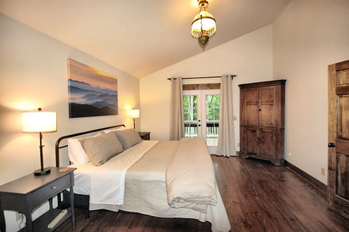 A peaceful queen guest bedroom with a comfortable bed dressed in high-end linens, nightstands with lamps for ambient lighting, a wardrobe, and a landscape painting above the headboard. Sunlight filters softly through a window or doorway, creating a restful, welcoming atmosphere.