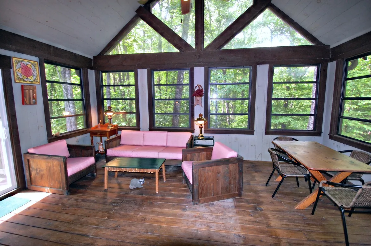 A sun porch lined with pink sofas and a wooden table, surrounded by large windows that let in dappled sunlight. The space is bright, cheerful, and perfect for quiet reading, watching sunsets, or sharing a story with friends and family.