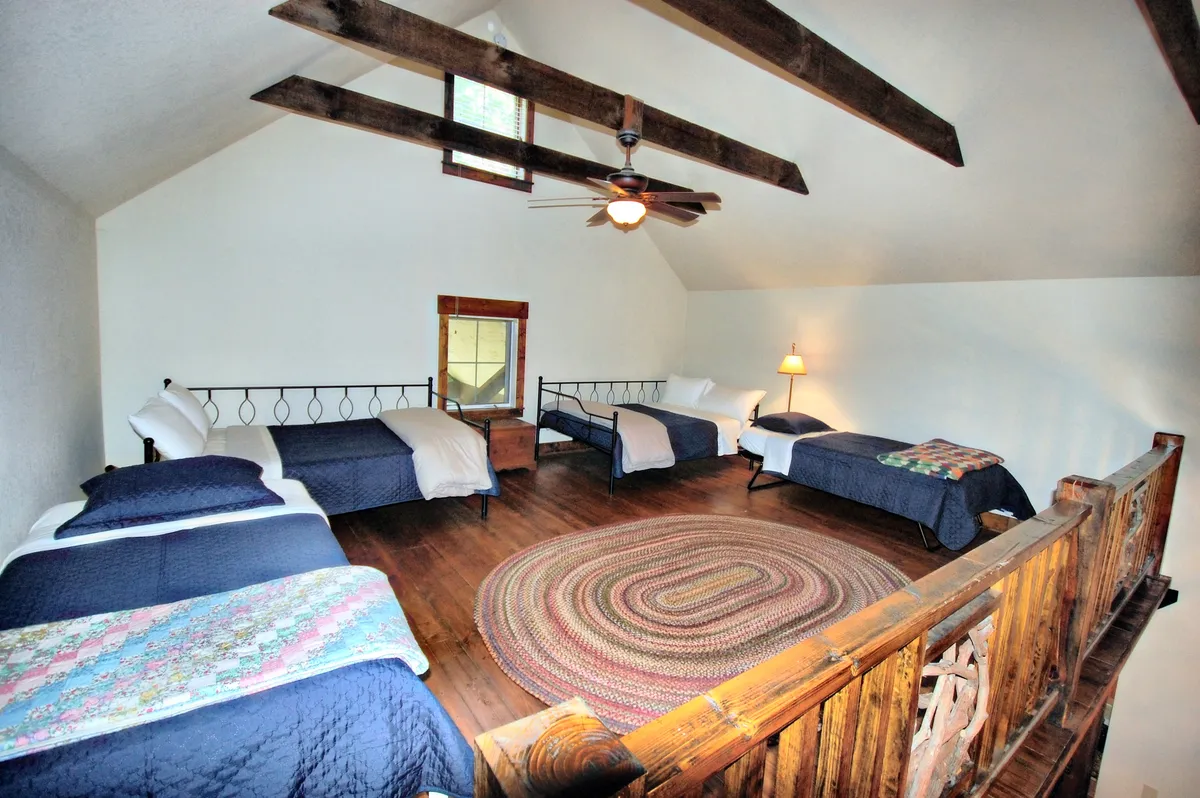 A cozy sleeping loft featuring four neatly made beds—two doubles and two twins—arranged under charming wooden beams. A round braided rug anchors the space, suggesting warmth and togetherness, while soft lighting and natural textures invite relaxation and late-night chats.