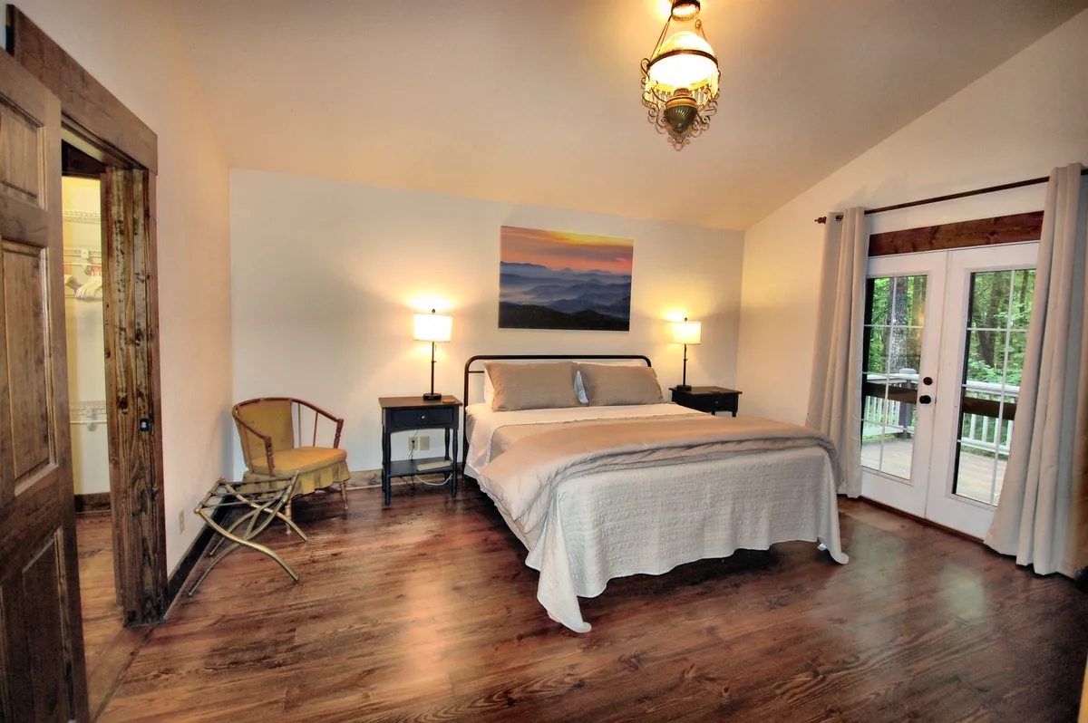 A serene king bedroom suite featuring a plush bed with a stylish headboard, coordinated bedding, and bedside tables with glowing lamps. Large French doors allow natural light to stream in, offering direct access to a private deck and the possibility of enjoying peaceful mornings outdoors. Elegant wall art and soft textures complete the tranquil ambiance.