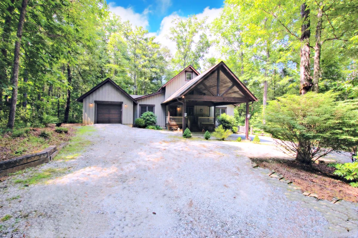 A wide exterior shot featuring the front porch, garage, and a backdrop of tall, mature trees. The tranquil setting showcases how the home is nestled within nature, making it an ideal base for outdoor activities. The spacious surroundings and visible tree line suggest opportunities for both relaxation and adventure in the immediate vicinity.