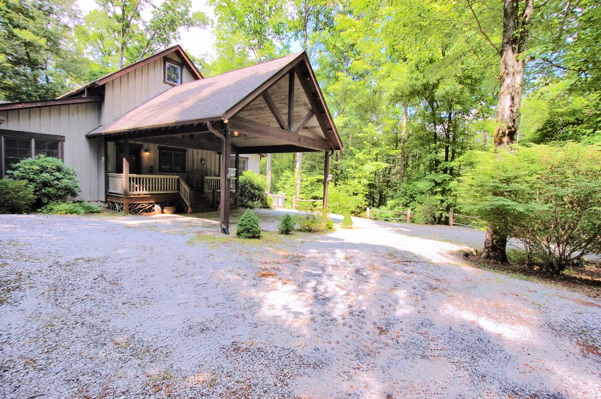 A cozy front porch with rustic wooden beams overlooks a backdrop of dense greenery. The scene highlights the connection between the home's comfort and the natural world beyond, with hints of trails and pathways leading away from the property into the woods. The greenery and porch setting suggest a peaceful spot for observing wildlife or gearing up for a family hike.